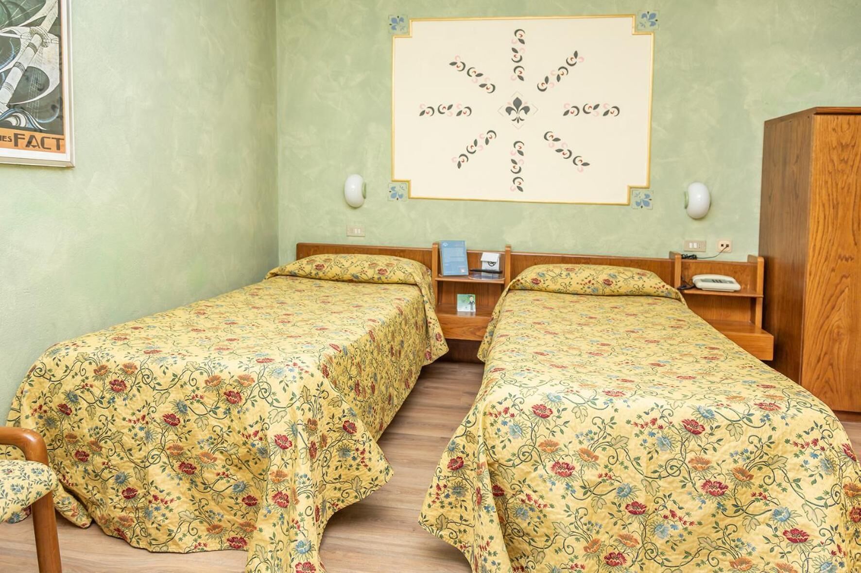 Una stanza con due letti singoli, un armadio in legno, un comodino con una lampada e un quadro appeso al muro.