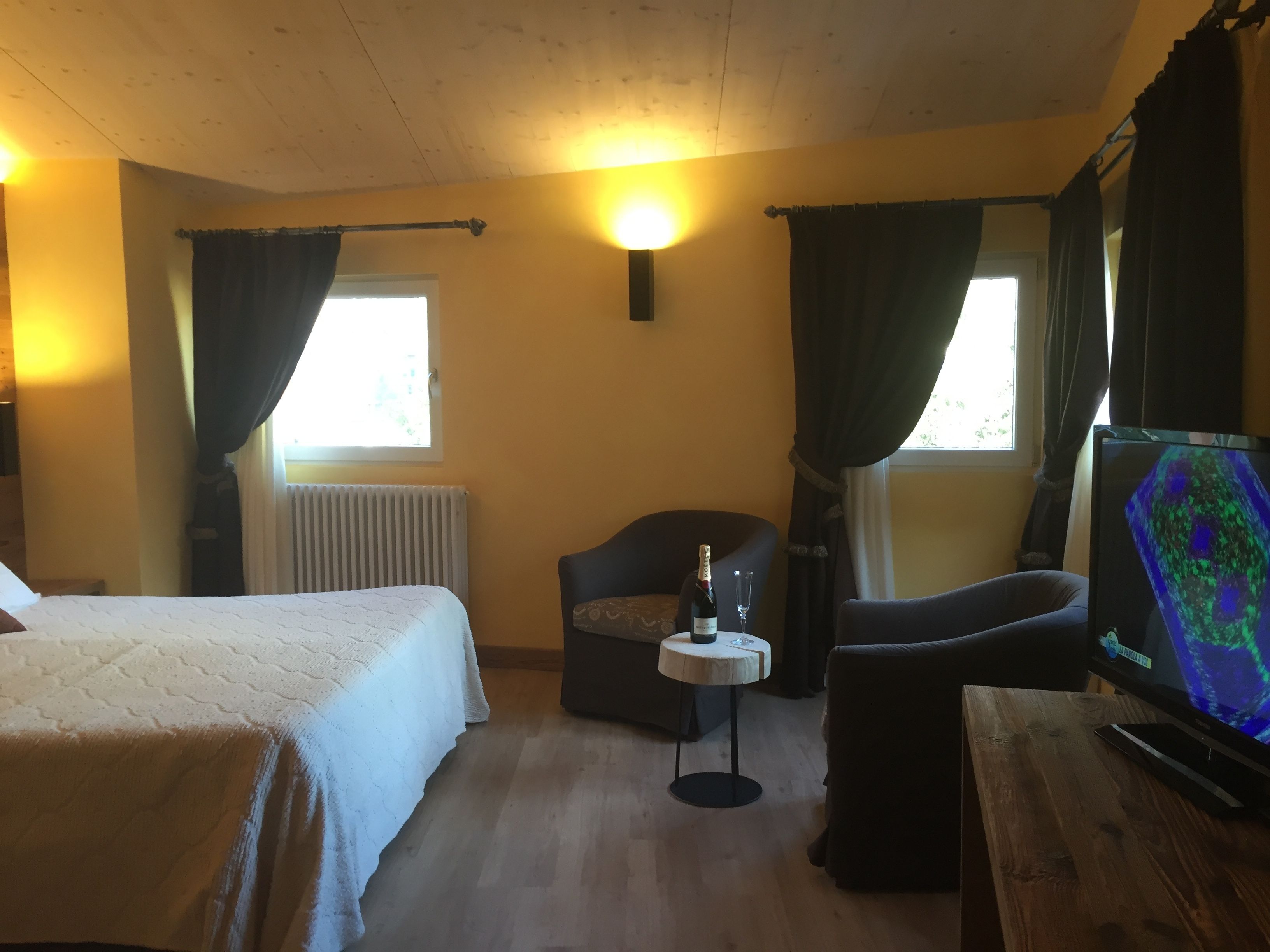 Ein Zimmer mit zwei Betten, einem kleinen Tisch mit Flasche und Glas, einem Sessel, einem Fernseher und einem Fenster mit Vorhängen.