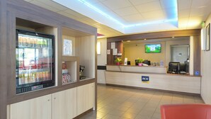 A hotel reception area with a counter, a television screen, a refrigerator, and a seating area. - ACE Hôtel Paris Roissy (Mitry-Mory)