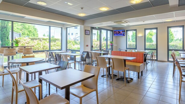 A spacious indoor dining area with multiple tables and chairs, large windows, and a television screen on the wall. - ACE Hôtel Paris Roissy (Mitry-Mory)