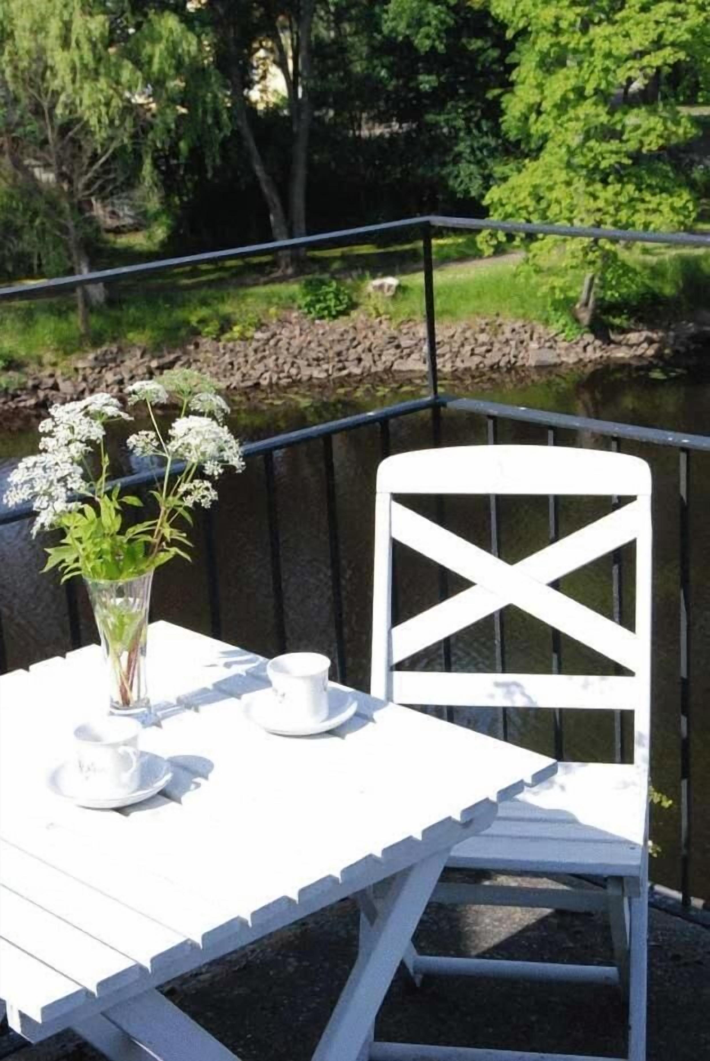 Una mesa blanca al aire libre con una silla, un florero con flores y un juego de taza y platillo.