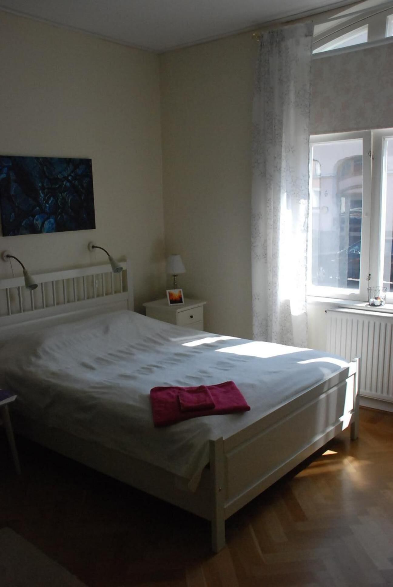 Un dormitorio con una cama grande, una mesita de noche con lámpara, un marco de fotos y una ventana con cortinas.