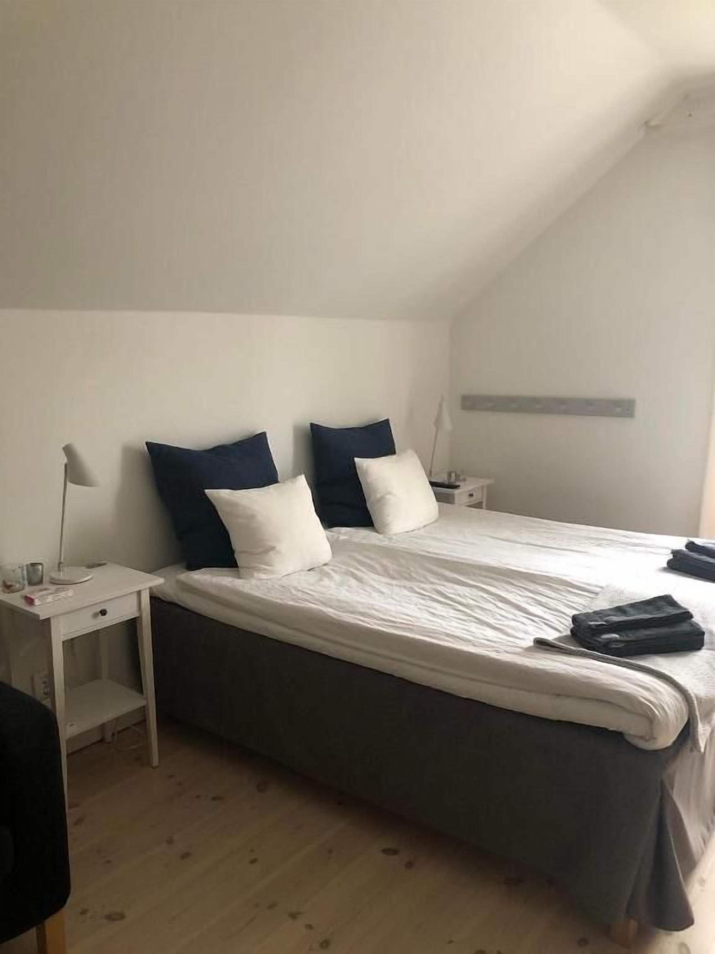Un letto singolo con materasso grigio e biancheria da letto bianca, due comodini con lampade e una parete bianca con ripiano.