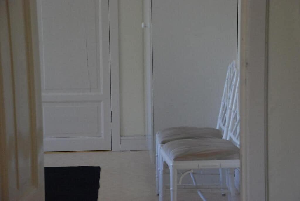 Un couloir étroit avec une porte blanche et une chaise blanche dotée d’un coussin.