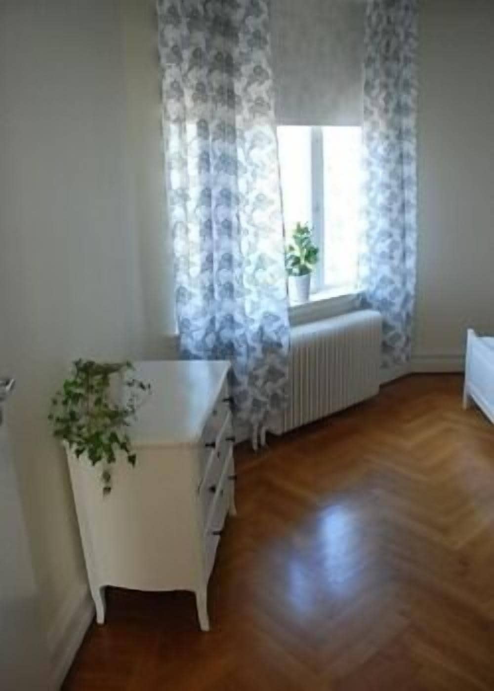 A room with a white desk, a radiator, and a window with patterned curtains.