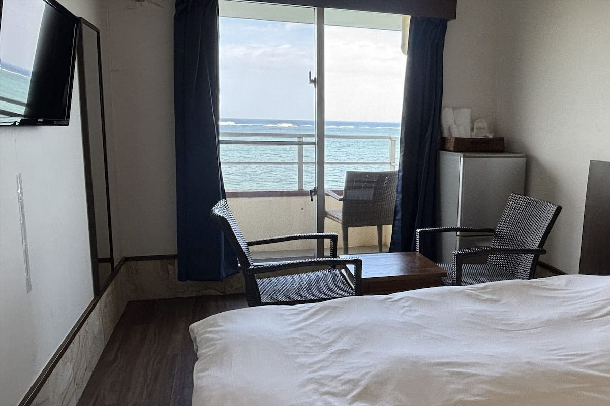 A hotel room with a bed, a desk, two chairs, a television, and a view of the ocean.