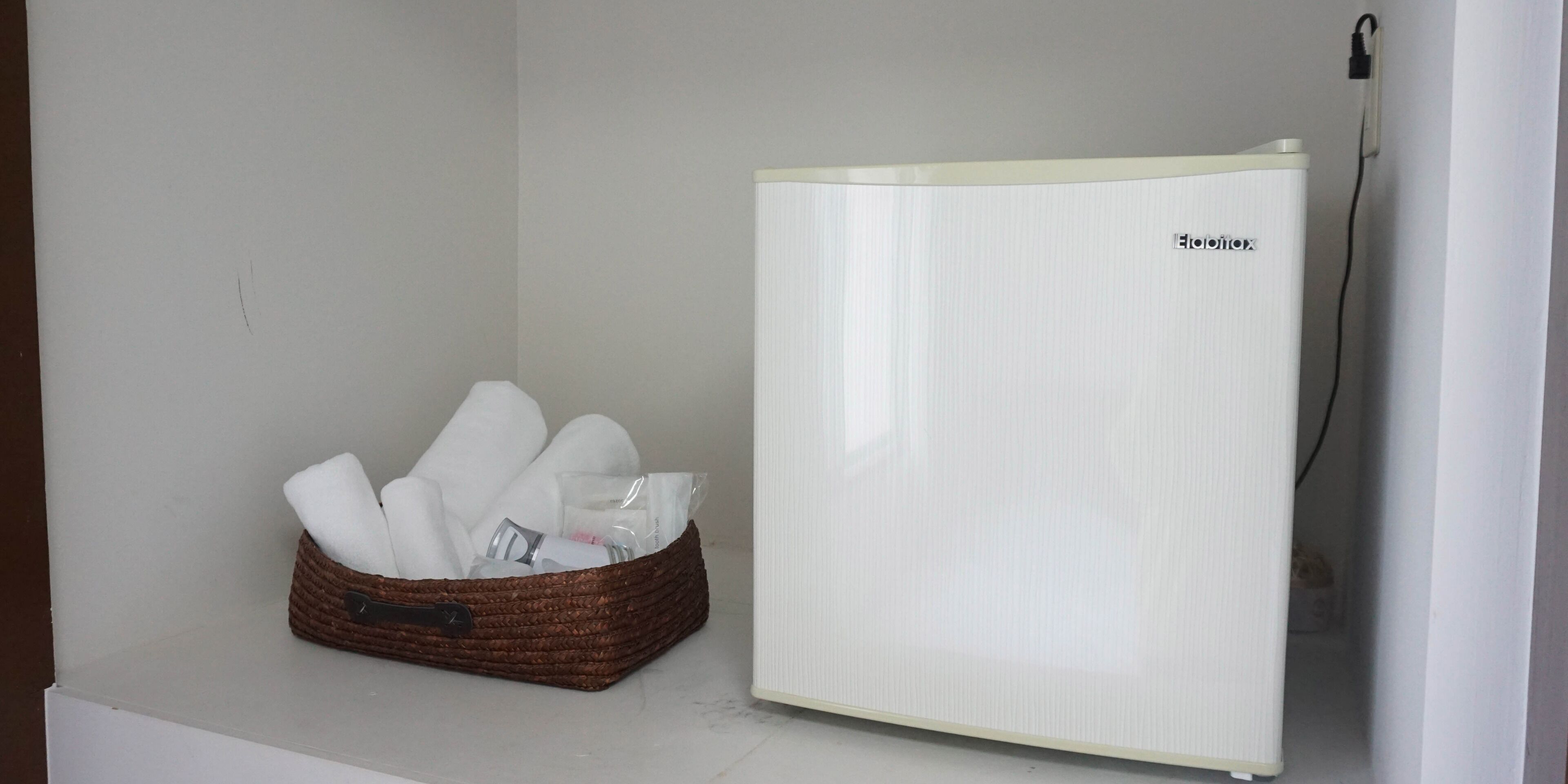 A white refrigerator with a brand name, a wicker basket with towels, and a power cord plugged in.