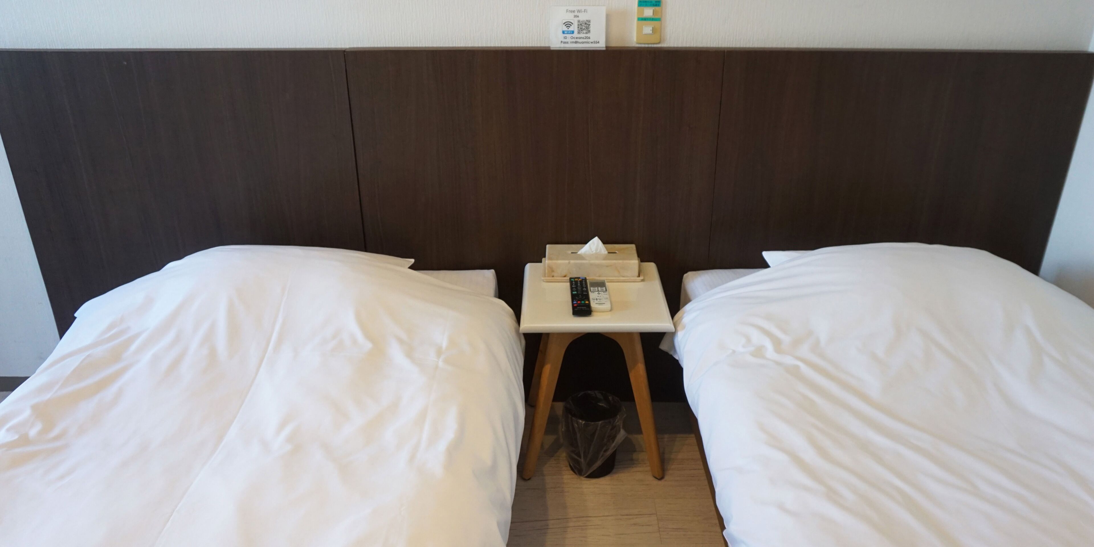 Ein Hotelzimmer mit zwei Betten, einem kleinen Tisch mit Fernbedienung, einem Mülleimer und einem Wandregal mit Annehmlichkeiten.