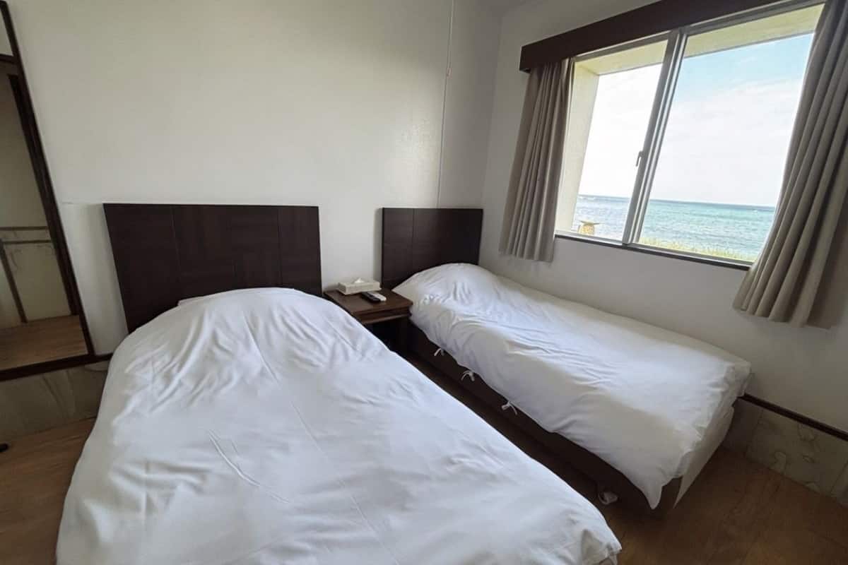 A hotel room with two single beds, a bedside table with a remote control, and a window with curtains.
