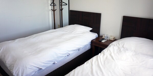 A hotel room with two single beds, a wooden headboard, a nightstand with a remote control, and a coat rack.