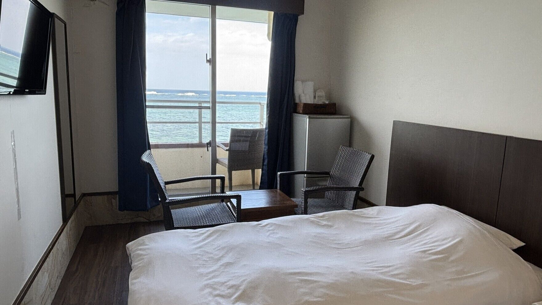 A hotel room with a bed, two chairs, a small table, a TV, and a view of the ocean.
