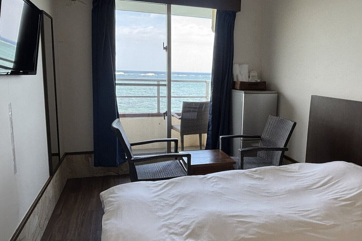 A hotel room with a bed, two chairs, a small table, a TV, and a view of the ocean.