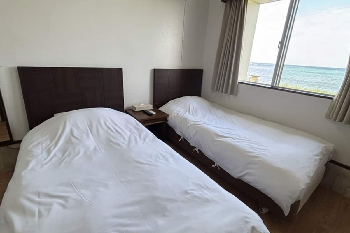 A hotel room with two single beds, a nightstand, a remote control, and a window with a view of the sea.