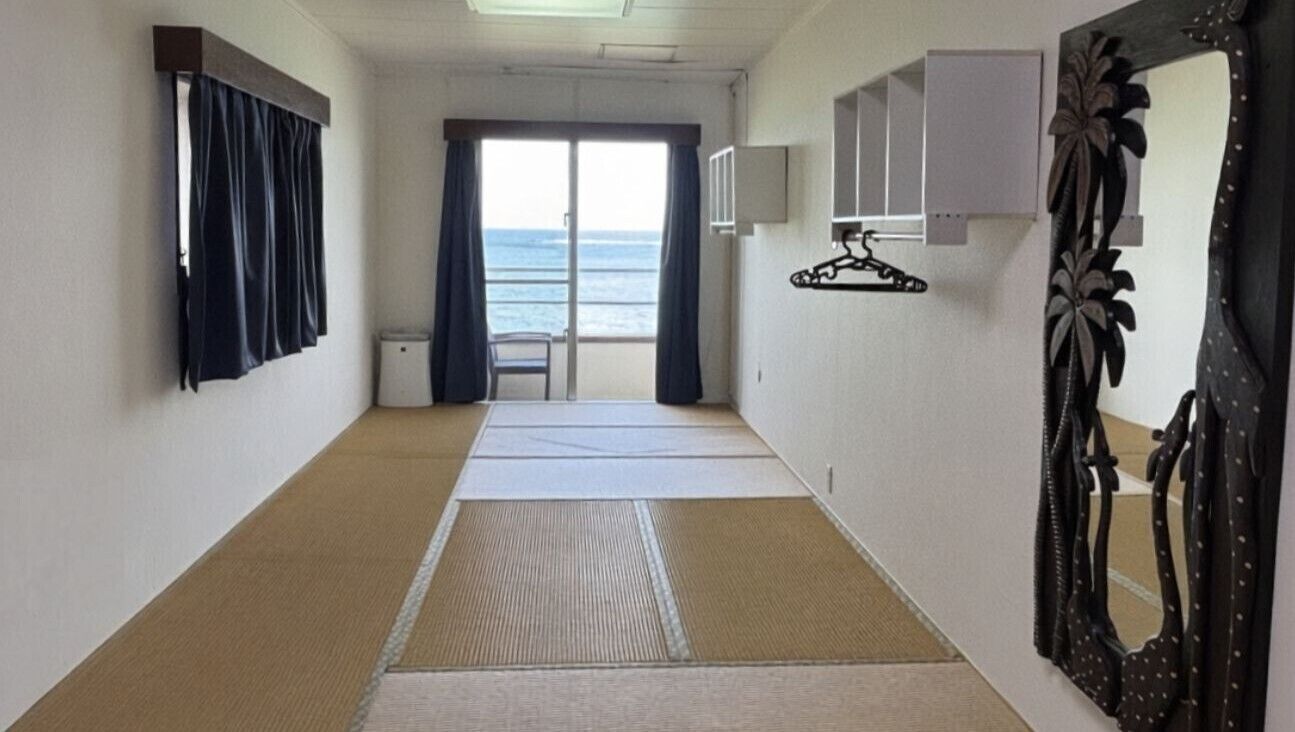 Een gang met tatami matten, een schuifdeur naar een balkon met uitzicht op zee, een stoel en een spiegel met een decoratieve lijst.