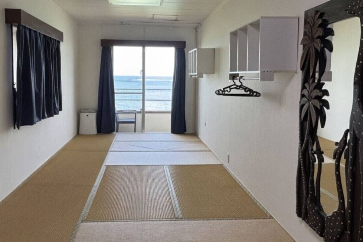 Een gang met tatami matten, een schuifdeur naar een balkon met uitzicht op zee, een stoel en een spiegel met een decoratieve lijst.
