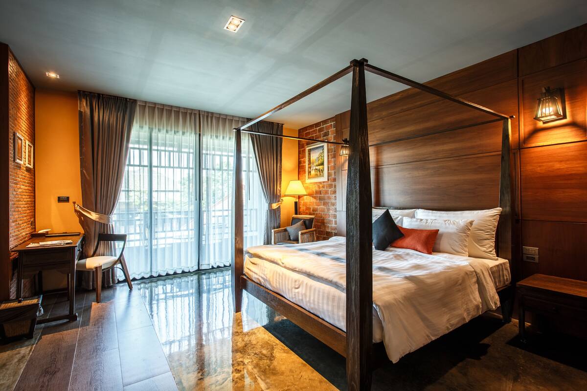 A four-poster bed with a wooden headboard, a desk with a chair, a lamp, and a view of the outdoors through large windows.