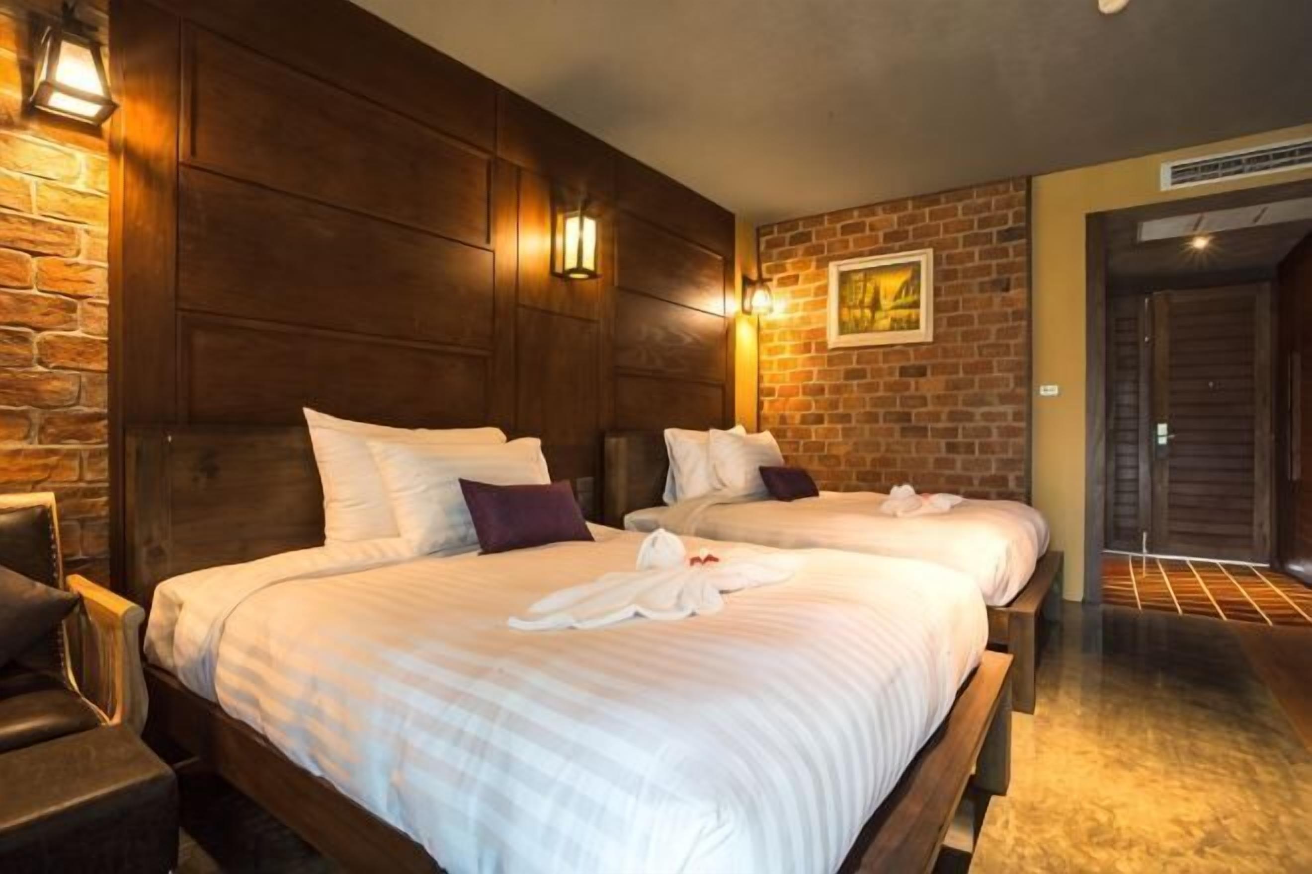 A hotel room with two beds, wooden headboards, a brick accent wall, and a door leading to the hallway.