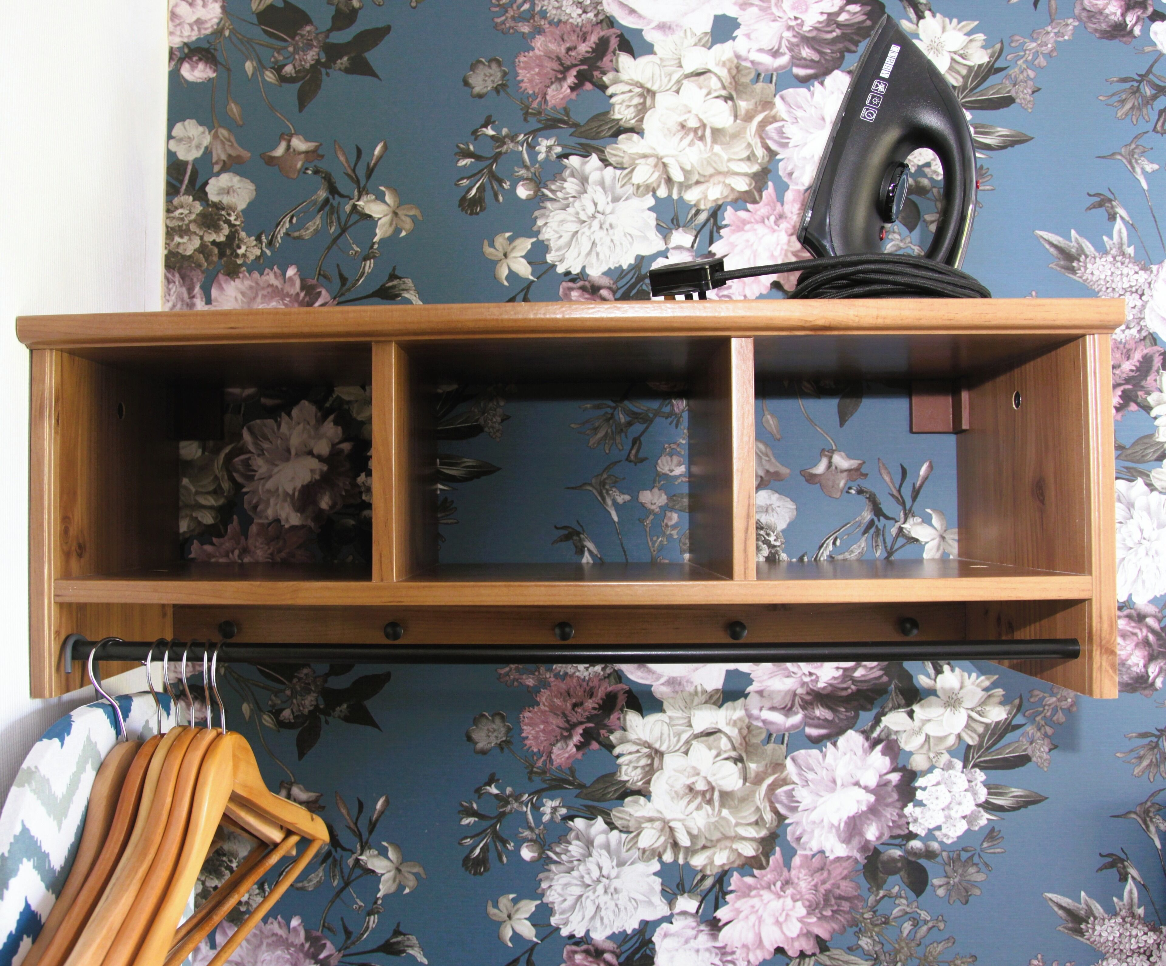 A wooden shelf with a floral wallpaper background, a hairdryer, and wooden hangers.