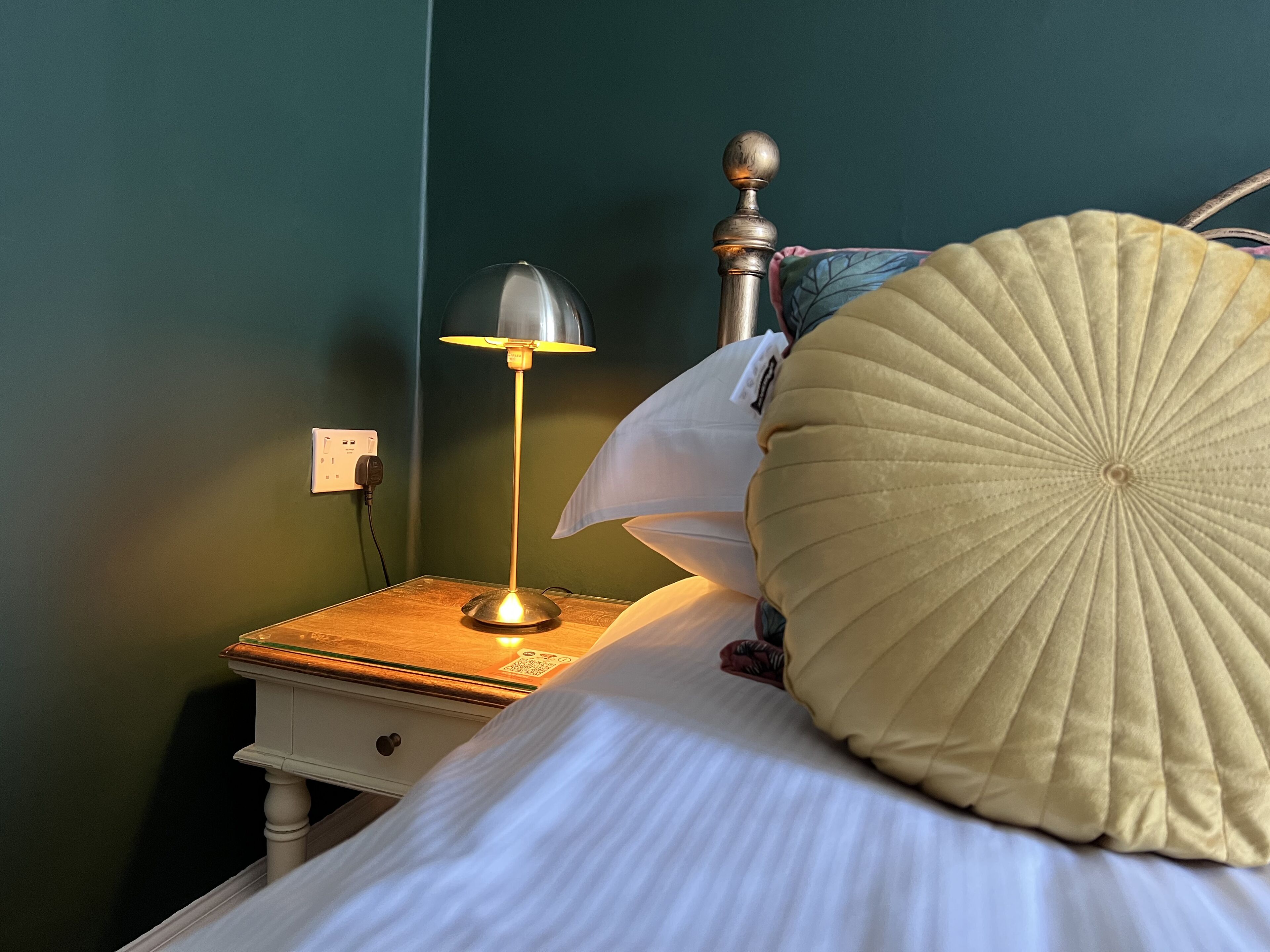 A neatly made bed with a white pillow and a beige decorative pillow, a bedside lamp on a wooden nightstand, and a green wall.