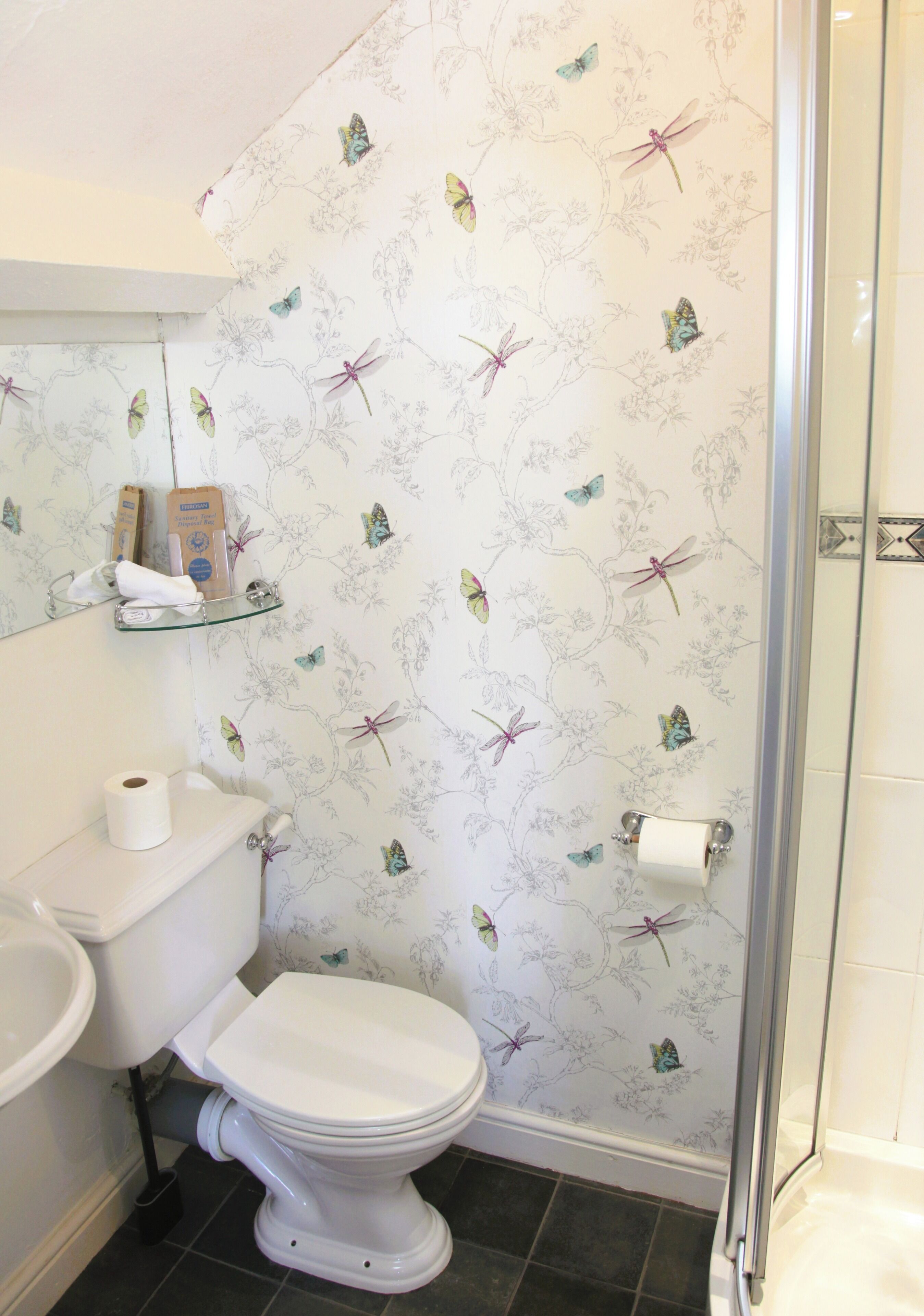 Un baño con inodoro, un estante de vidrio con artículos de aseo y una zona de ducha.