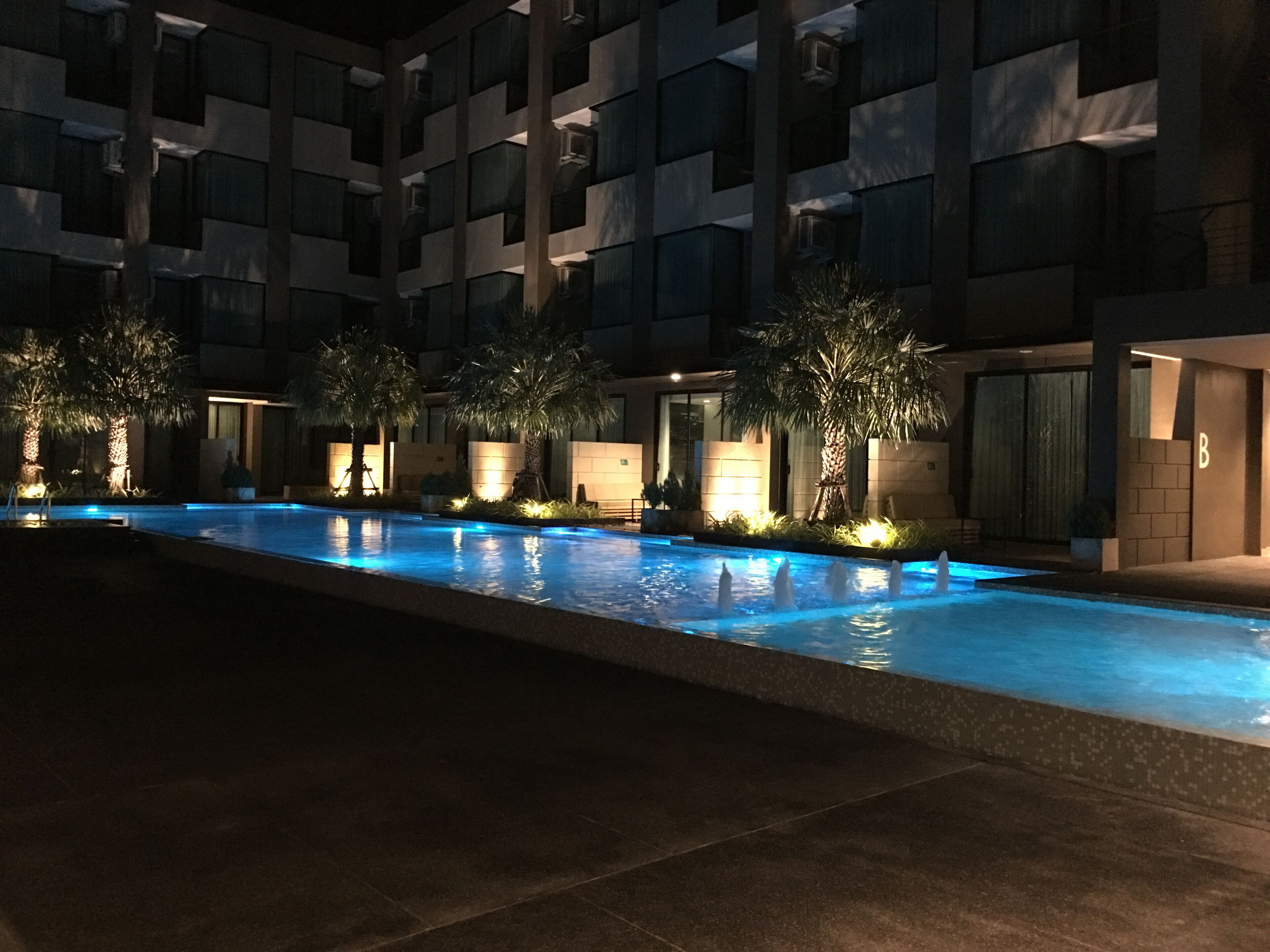 Una piscina nocturna con bordes iluminados, rodeada de palmeras y un edificio de varios pisos con balcones.