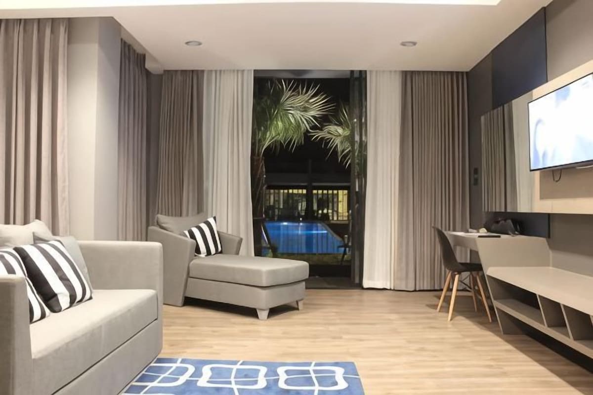 A modern living room with a sofa, armchair, and a dining table. A large TV is mounted on the wall.