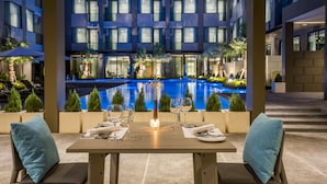 Comedor con una mesa dispuesta para dos, con vista a una piscina y un edificio de múltiples ventanales.