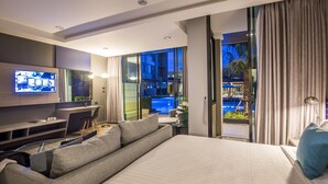 Una habitación de hotel moderna con una cama grande, un televisor montado en la pared, un área de estar con sofá y sillones, y vistas a una piscina a través de amplios ventanales.