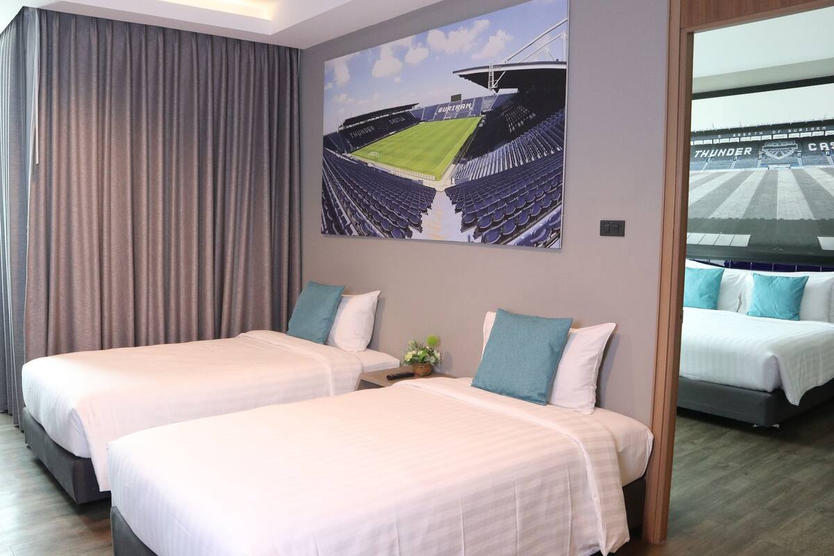 Une chambre d’hôtel avec deux lits, une grande fenêtre avec des rideaux, un tableau représentant un stade et une vue sur le stade depuis la fenêtre.
