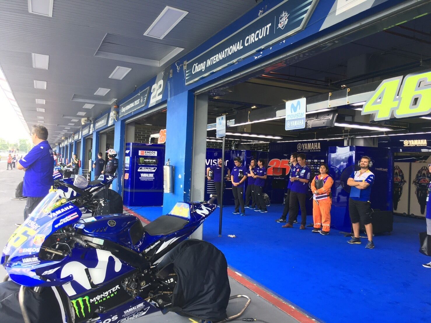 A motorcycle in a garage with a blue and white color scheme, a sign reading "Chang International Circuit", and several people standing in the background.