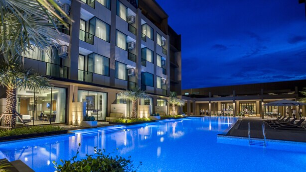 Un hotel moderno con una amplia piscina exterior, palmeras y una fachada bien iluminada.