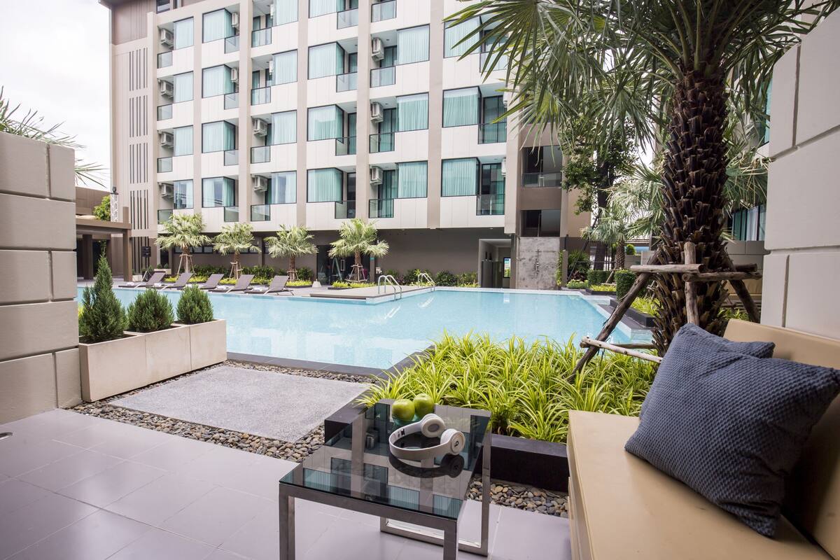 Un espace piscine moderne dans un hôtel, avec une table en verre, des chaises longues et un palmier.