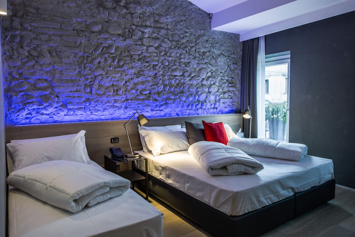 A modern hotel room with a stone wall, a large bed with white bedding, a bedside table with a lamp, and a view of the outdoors.