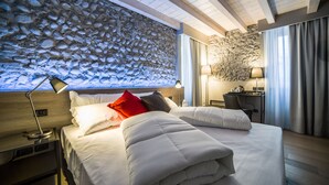A hotel room with a stone wall, a bed with white bedding, a desk with a lamp, and a view of the sea through a window. - Locanda al Vescovo (Verona)