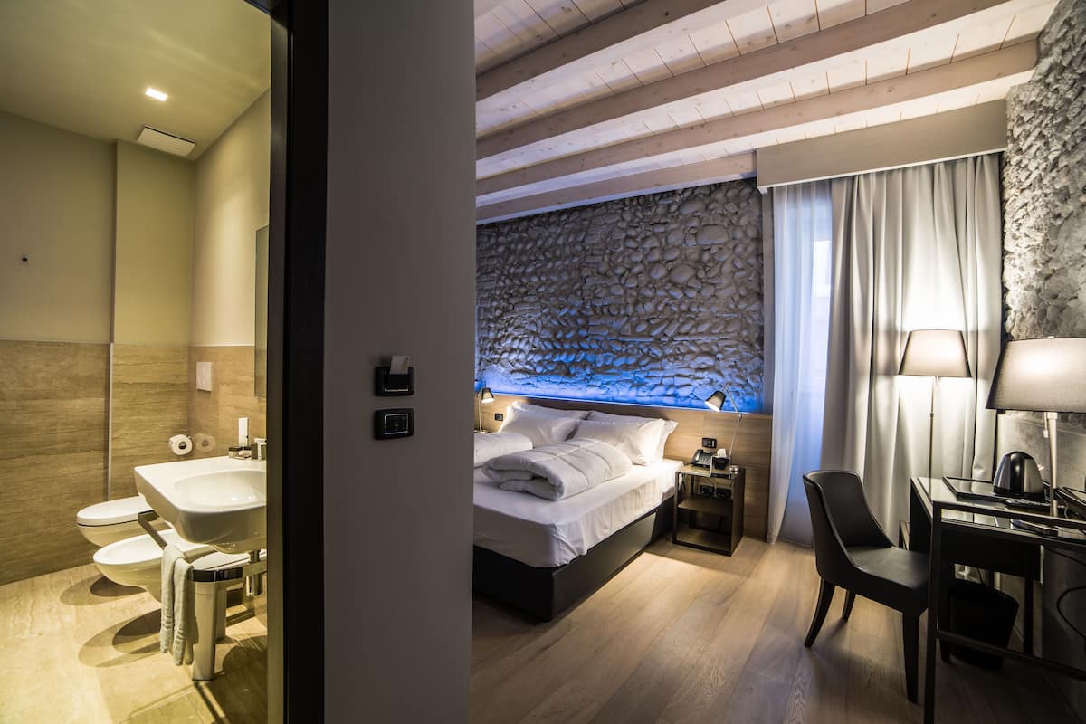A modern hotel room with a stone accent wall, a bed, a desk, and a bathroom with a sink and toilet.