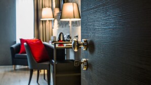 A hotel room with a wooden door, a desk with a lamp, a red chair, and a sofa with a red cushion. - Locanda al Vescovo (Verona)