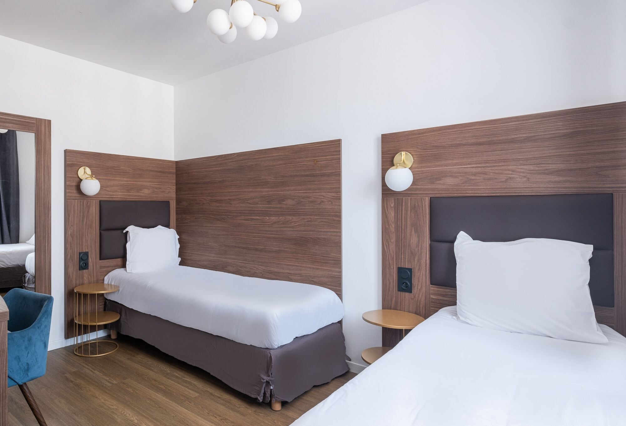 Una habitación de hotel moderna con dos camas individuales, una mesita redonda pequeña, una silla azul y suelo de madera.
