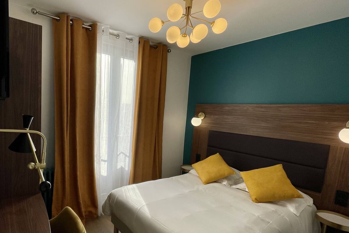 A hotel room with a large bed, two yellow pillows, a wooden headboard, a nightstand with a lamp, a chair, and a window with curtains.