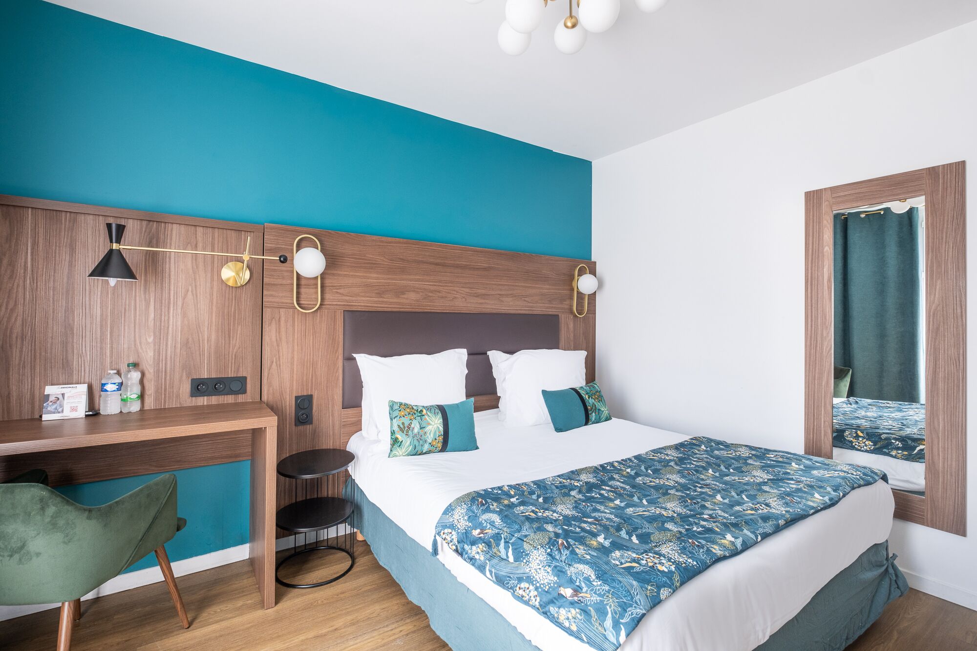 A modern hotel room with a teal accent wall, a wooden headboard, a desk with a chair, a bed with a floral bedspread, and a window with curtains.