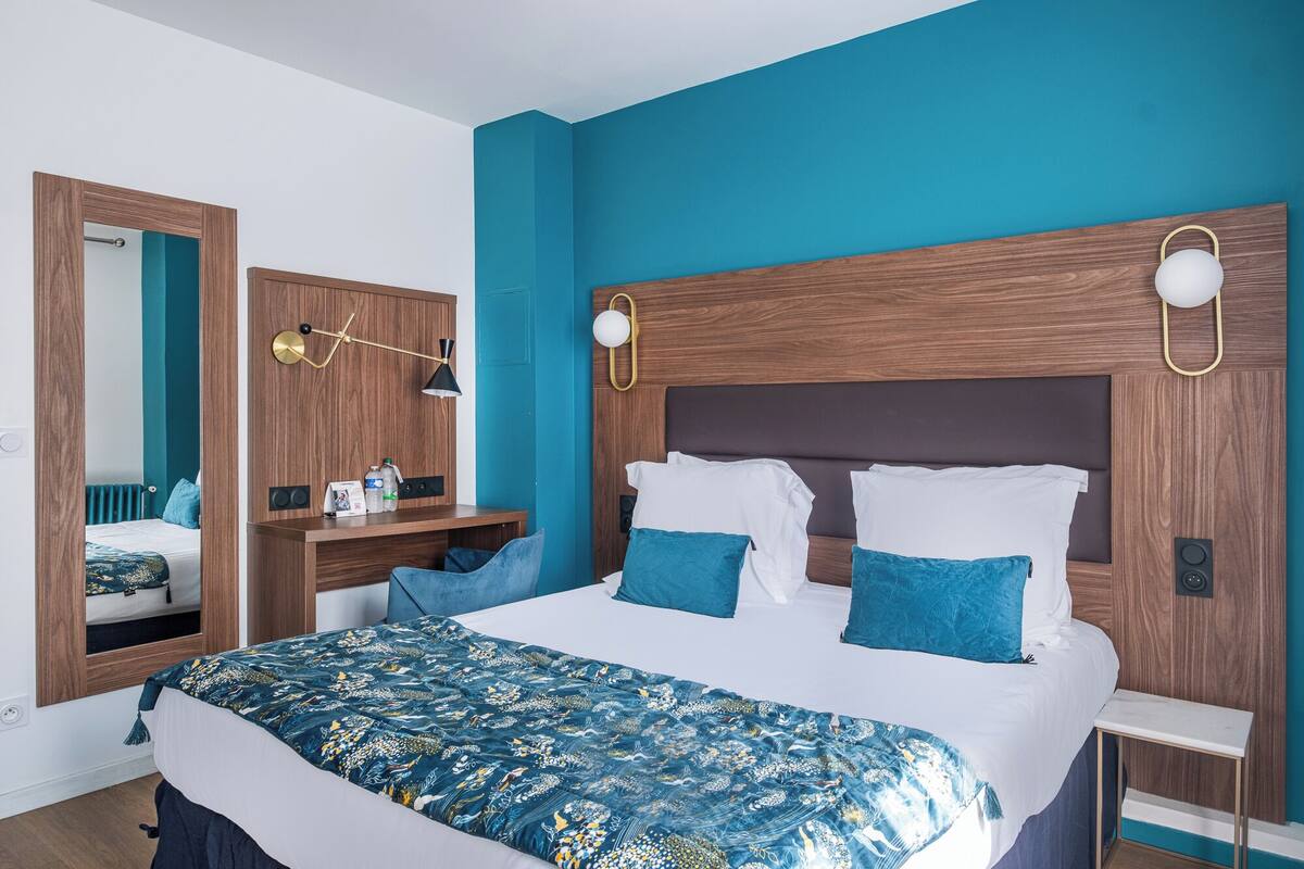 A modern bedroom with a large bed, blue walls, wooden headboard, and a bedside table with a lamp.