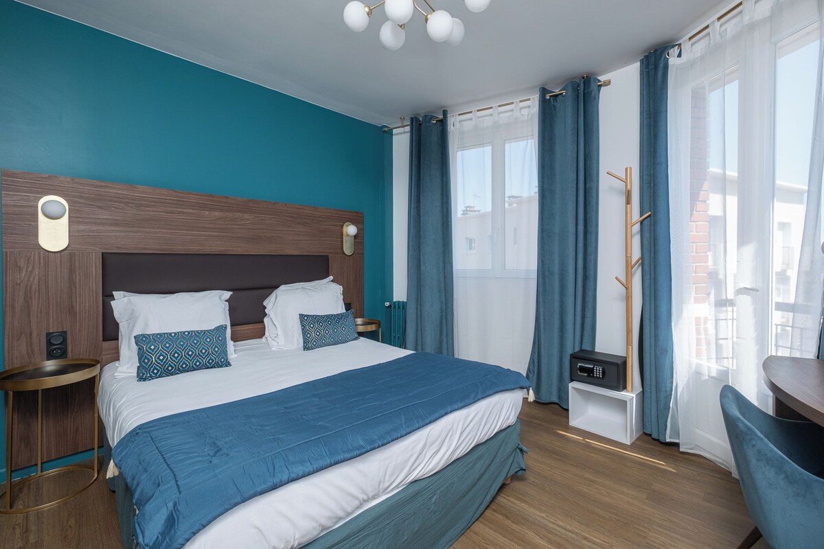 A modern hotel room with a large bed, a wooden headboard, a bedside table, a chair, and a view of the city.
