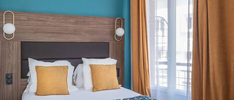 Una habitación de hotel con cama, dos almohadas, cabecera, mesita de noche, lámpara y ventana con cortinas.