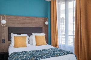 Quarto de hotel com cama, duas almofadas, cabeceira, mesa de cabeceira, abajur e janela com cortinas.