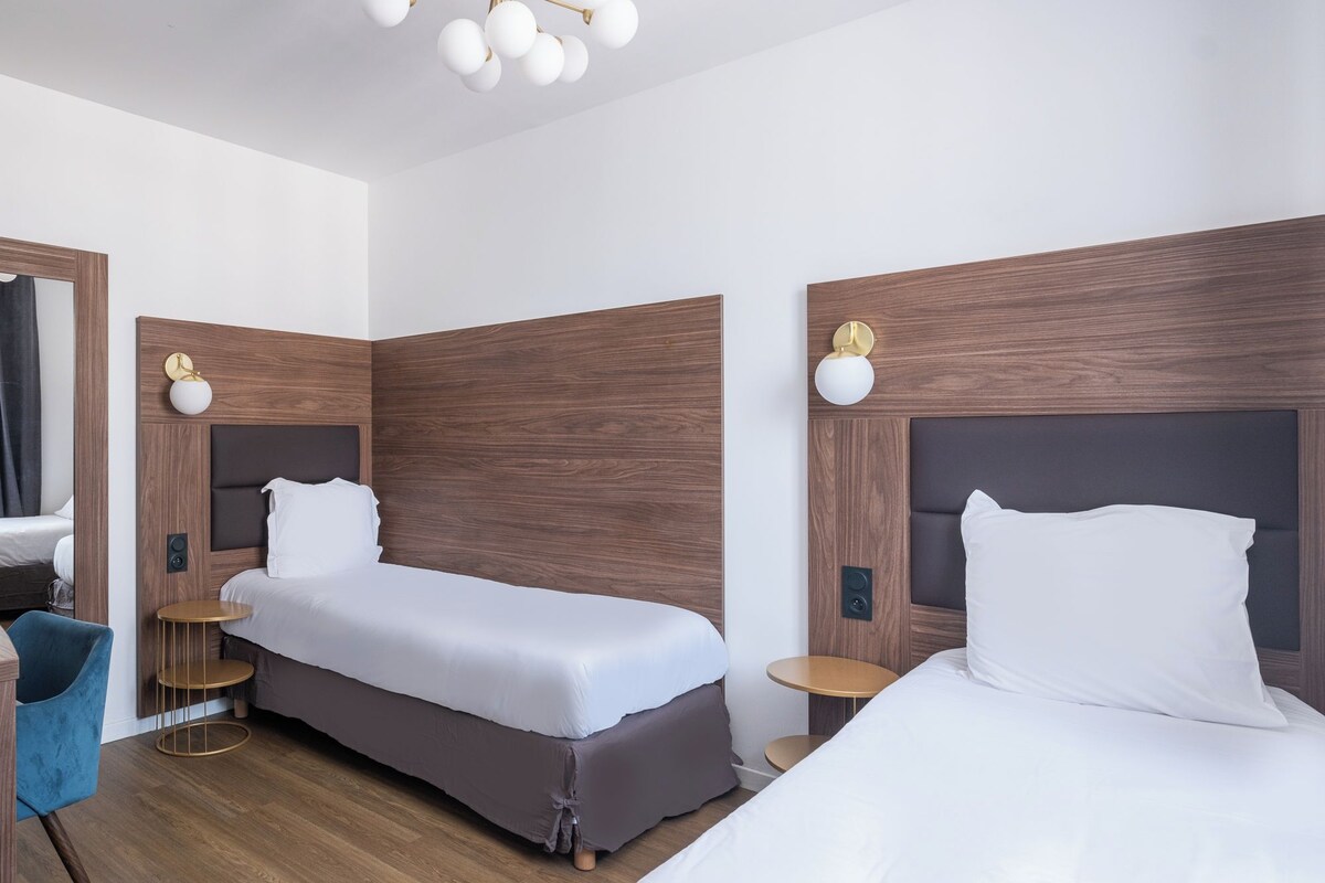 A modern hotel room with two single beds, a small round table, a blue chair, and a wooden floor.