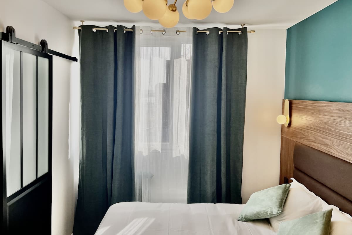 A hotel room with a bed, curtains, a ceiling light fixture, and a view of buildings outside.