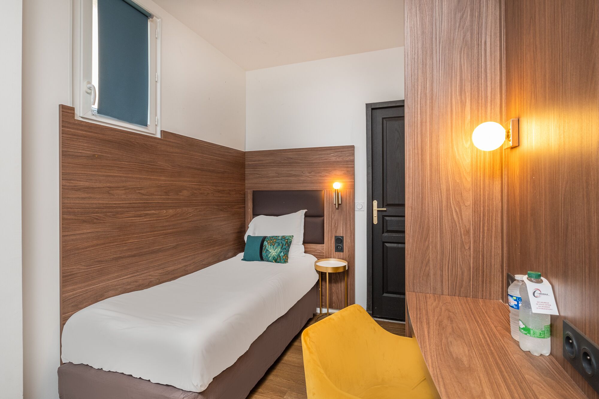 A compact hotel room with a single bed, a small bedside table, a chair, and a door.