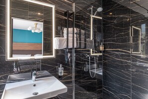 A modern bathroom with a marble wall, a white sink, a glass shower enclosure, and a framed artwork above the mirror.