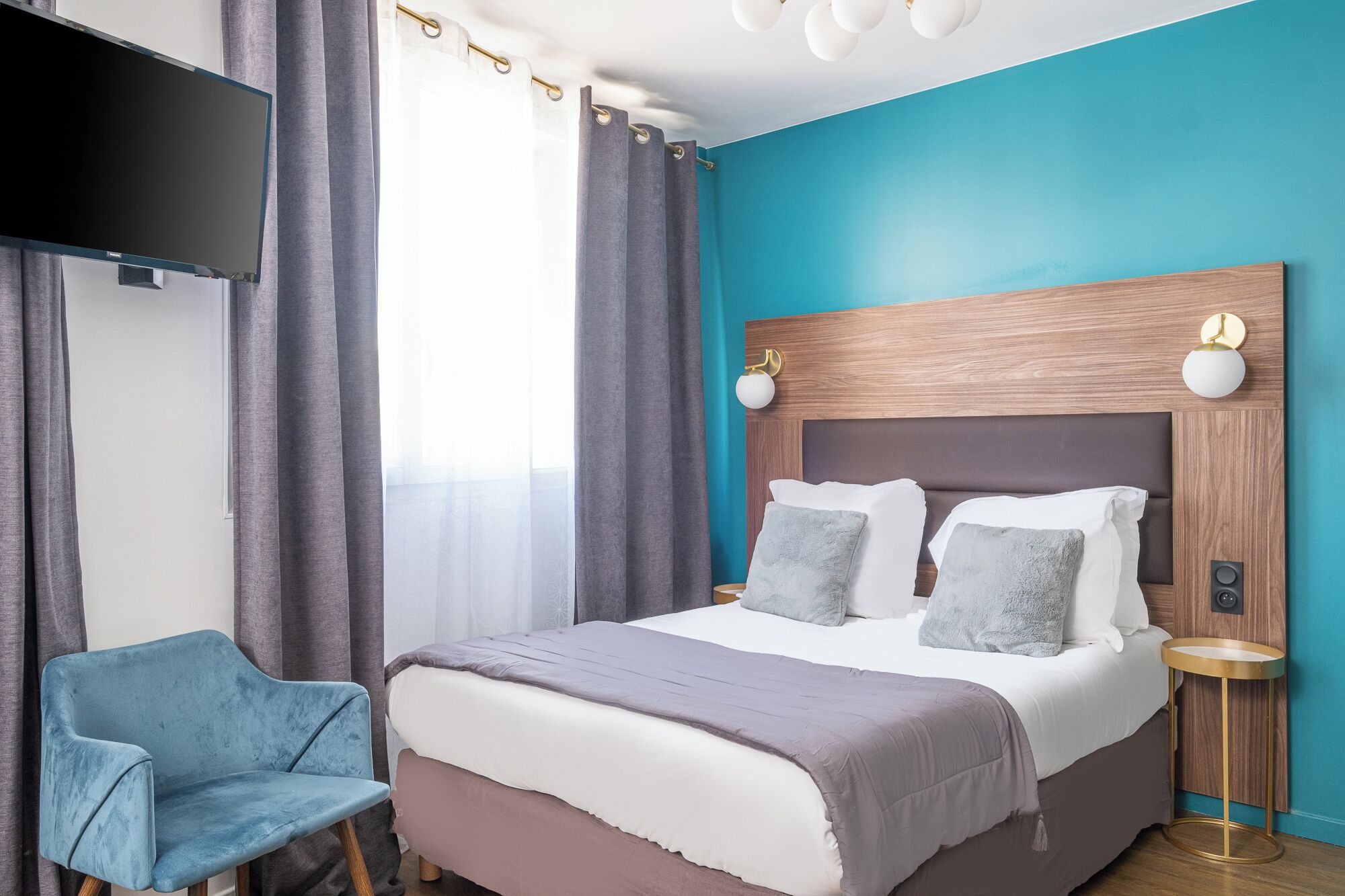 A modern hotel room with a large bed, a blue armchair, and a flat-screen TV mounted on the wall.