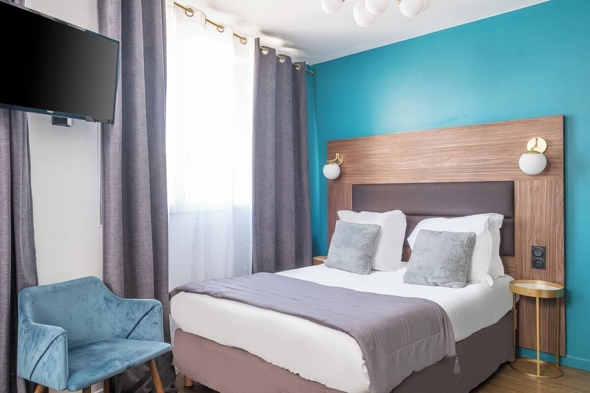 A modern hotel room with a large bed, a blue armchair, and a flat-screen TV mounted on the wall.