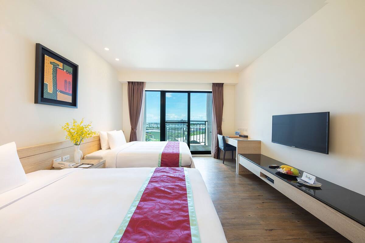 A hotel room with two beds, a desk, a TV, and a balcony with a view.